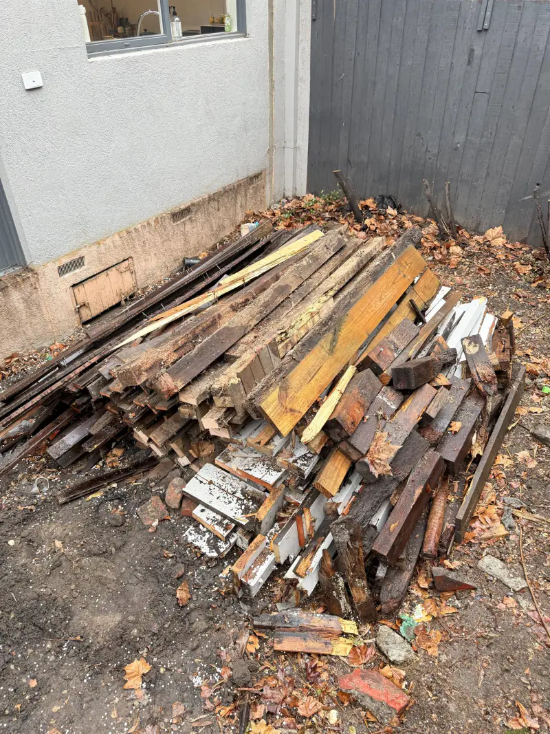 Pile of old decking timber, framing offcuts and painted architraves stacked against a house wall before removal
