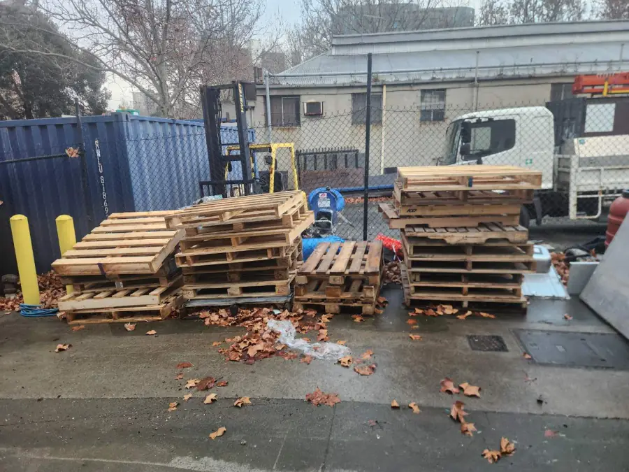 Four stacks of timber pallets and loose packaging filling a commercial yard beside a shipping container