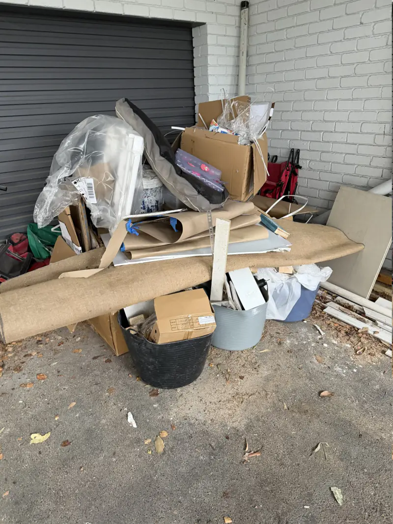 A garage entrance piled with flattened cardboard, a rolled carpet, hessian, boxes and household clutter